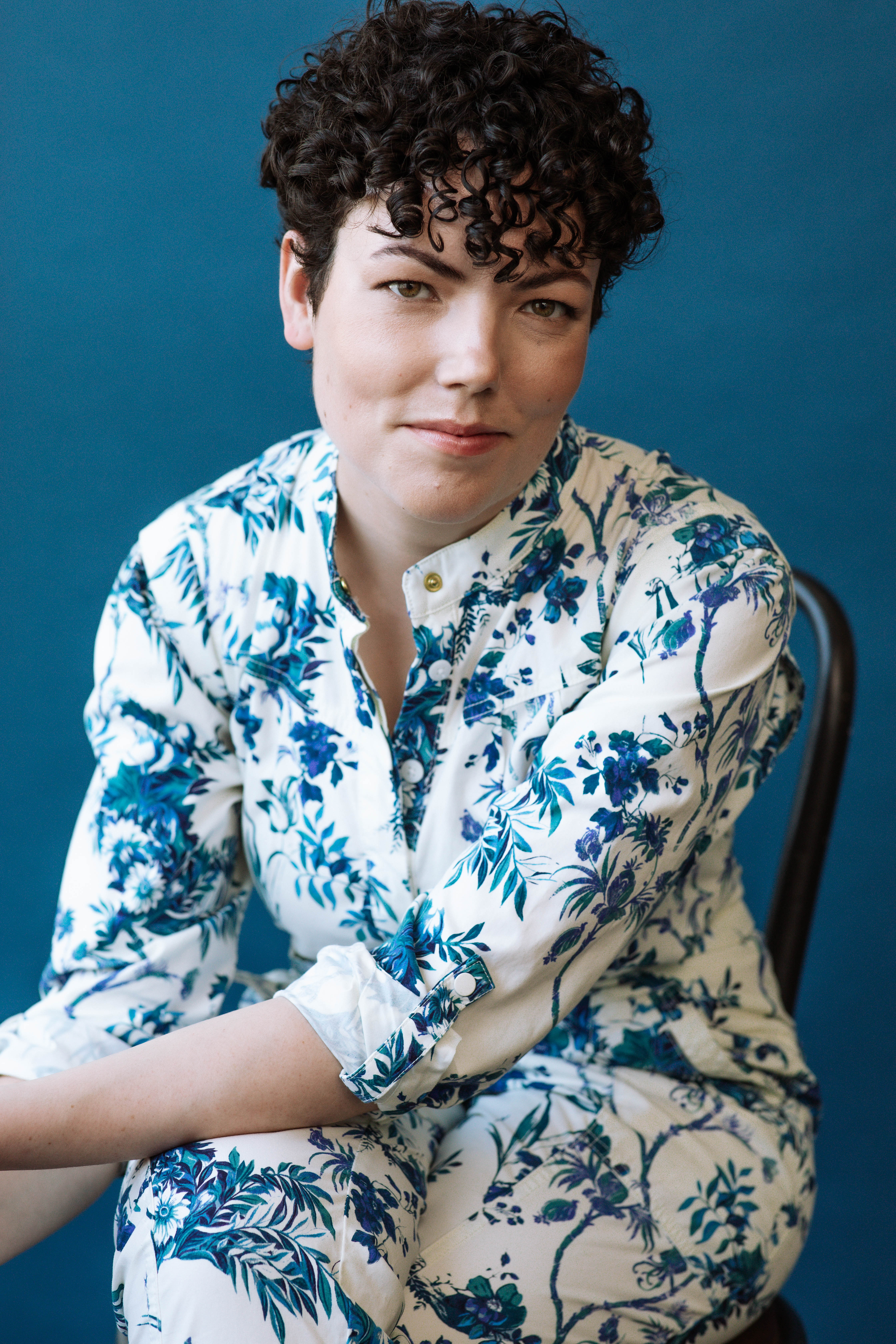 Bryn Kennedy in a white dress with blue flowers, sitting in a chair with hands clasped, against a blue backdrop
