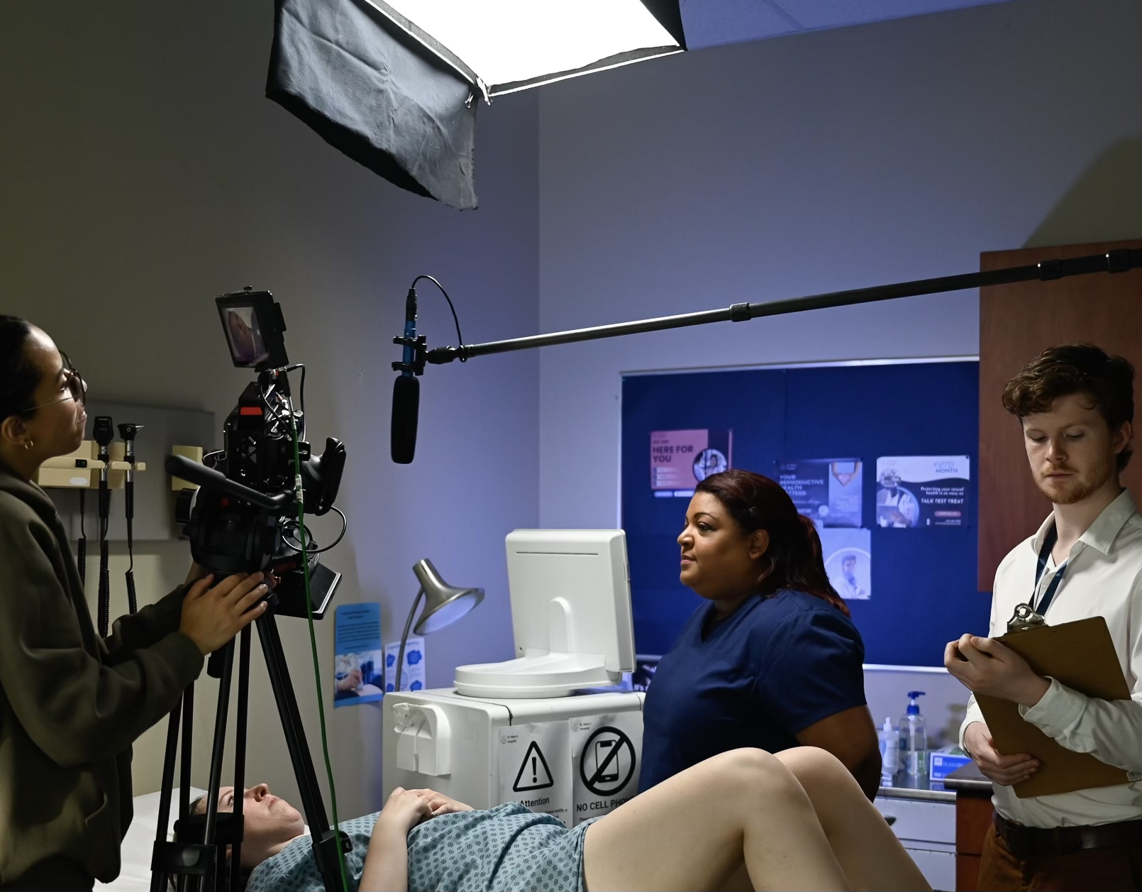Alice (Sarah Hime) is on an operating table in a doctor's room. The DP (Carine Zahner) is filming the nurse (Natasha Bromfield) and the student (Ben Caldwell), who are standing beside Alice.